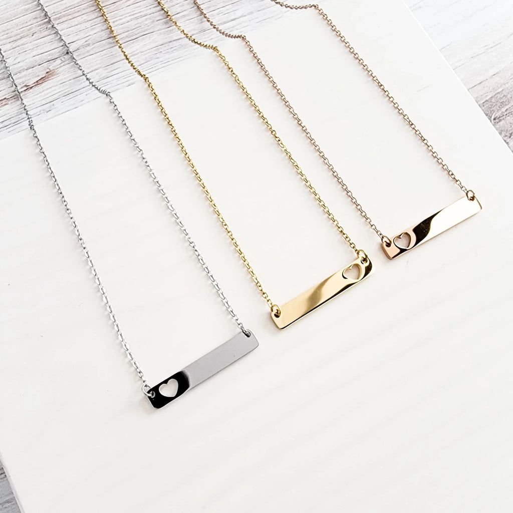 Roco Engraving custom bar necklaces in silver, gold, and rose gold stainless steel with heart cutouts on white wood.
