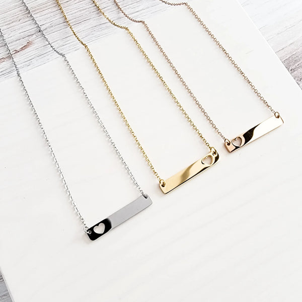 Roco Engraving custom bar necklaces in silver, gold, and rose gold stainless steel with heart cutouts on white wood.