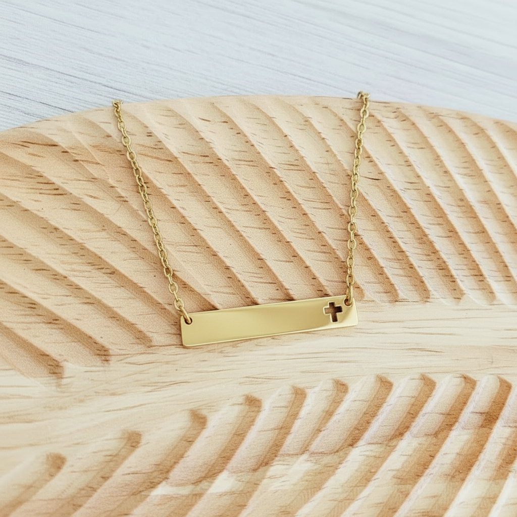 Roco Engraving gold stainless steel horizontal bar necklace with a cross cutout on a textured wooden display tray.
