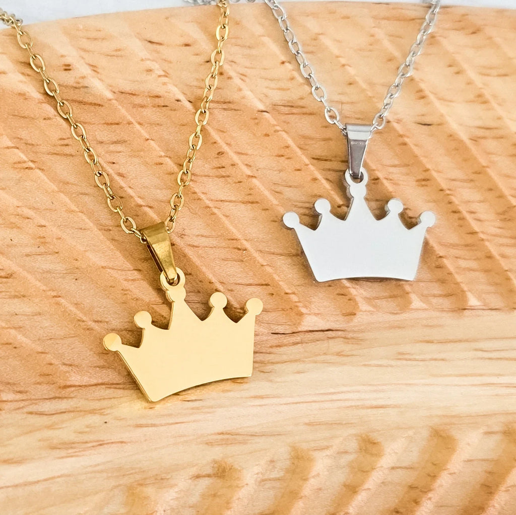 Roco Engraving gold and silver stainless steel crown pendants on chains resting on a wooden surface.