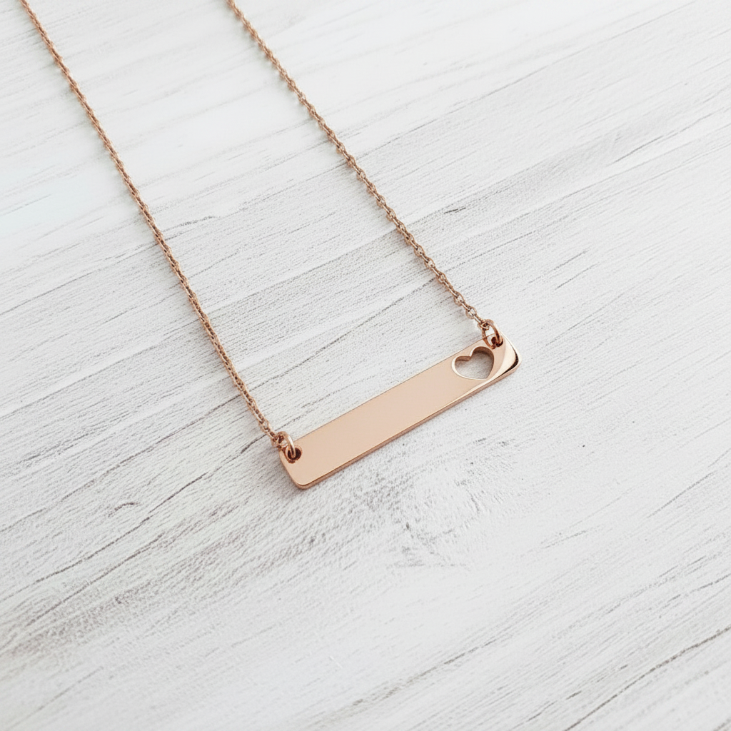 Roco Engraving rose gold bar necklace with a heart cutout on a delicate chain against a white wood background.